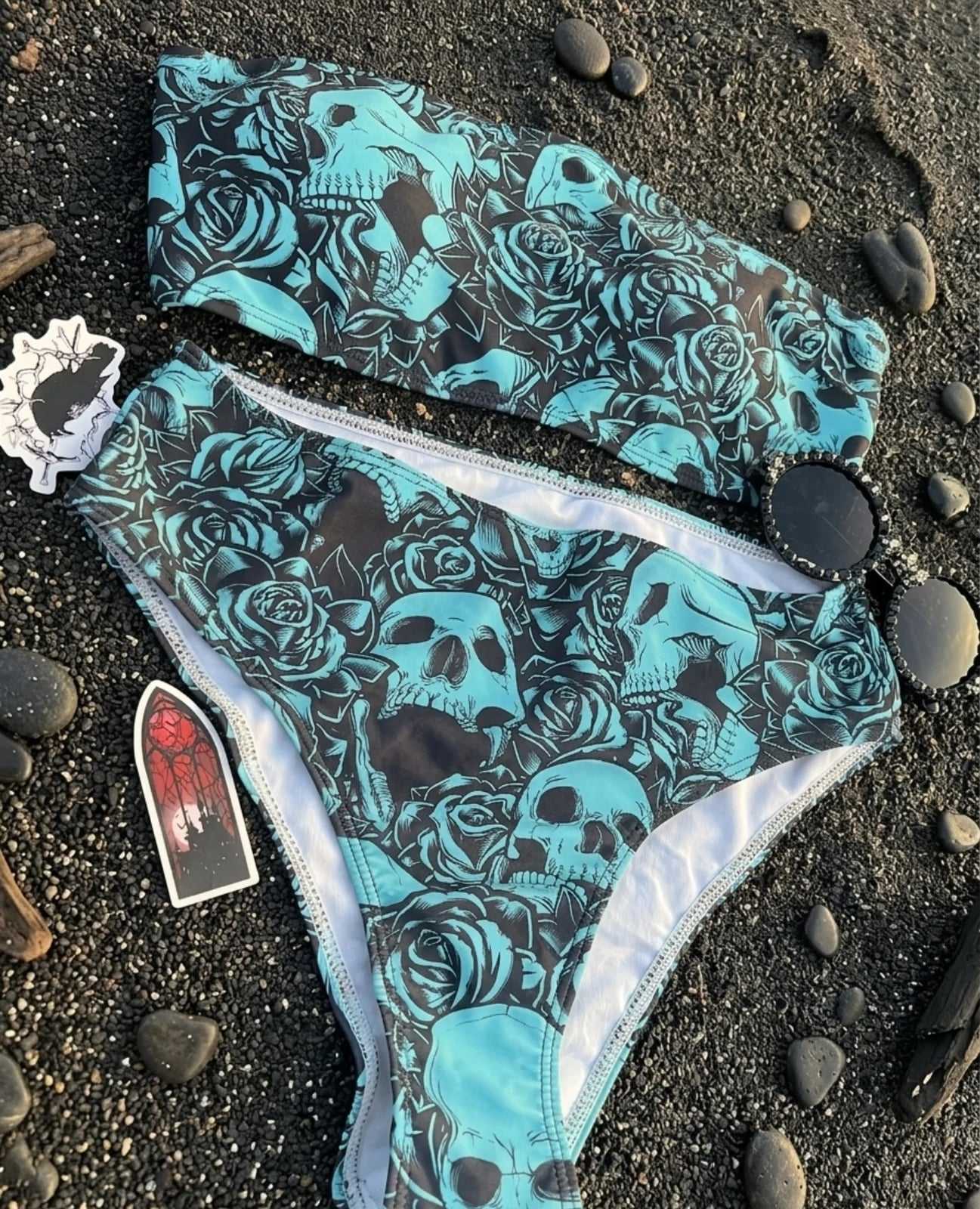 Bikini bandeau con estampado de calaveras y rosas en celeste y negro, junto a piedras, lentes y stickers sobre arena negra