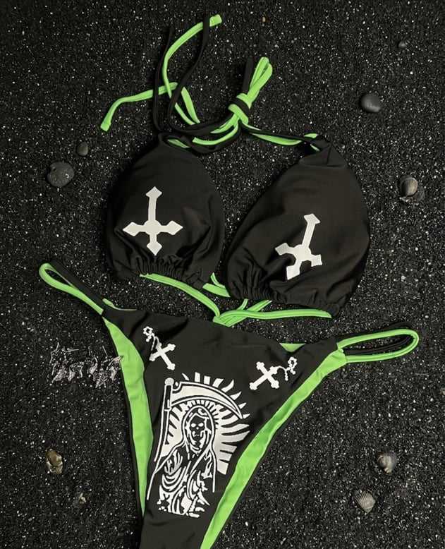 Bikini negro con ribete verde neón, cruces invertidas blancas y una parca estampada, sobre arena negra