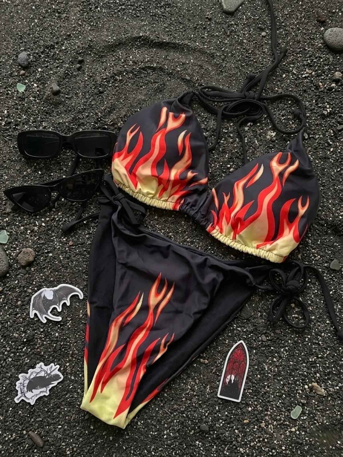 Bikini negro con estampado de llamas rojas y amarillas sobre arena volcánica negra, junto a lentes de sol negros y stickers góticos de murciélago y ataúd