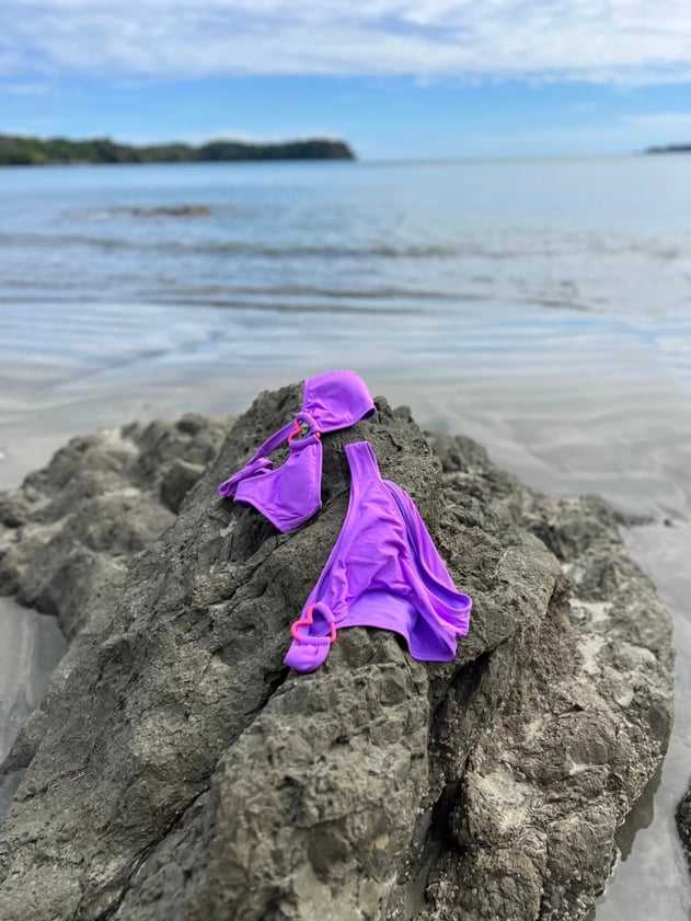 Bikini morado sobre una roca volcánica frente al mar, con una isla al fondo