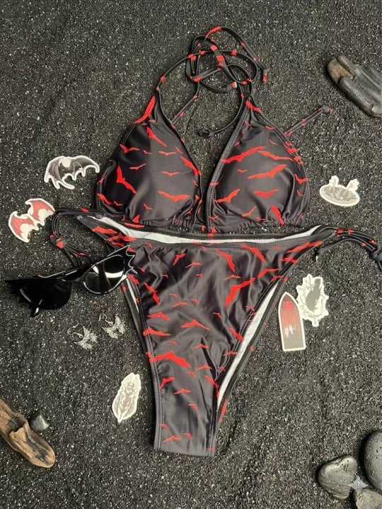 Bikini negro con estampado de murciélagos rojos, rodeado de stickers de murciélagos, lentes negros y madera sobre arena negra