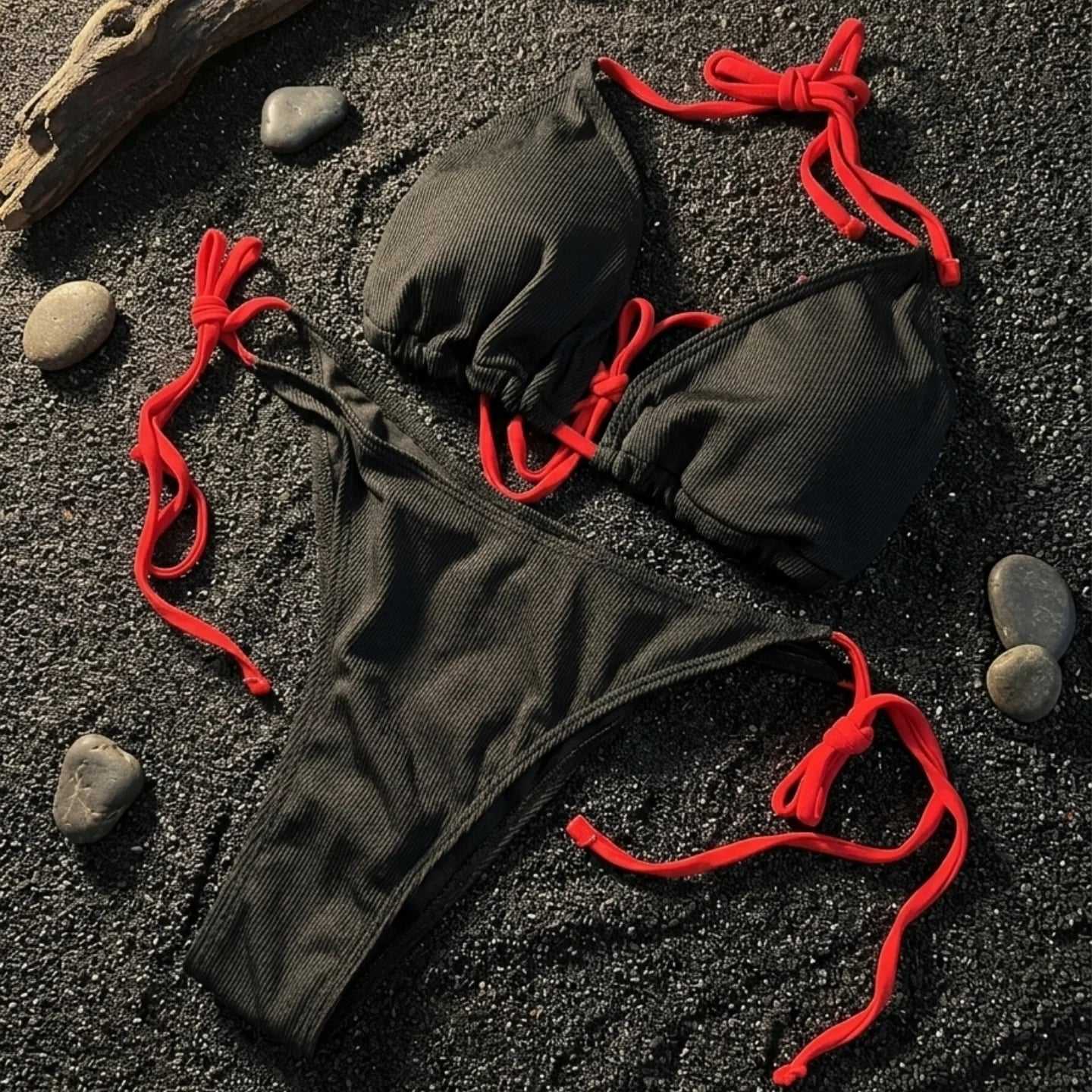 Bikini negro acanalado con tiras rojas para amarrar, sobre arena negra con piedras y un trozo de madera