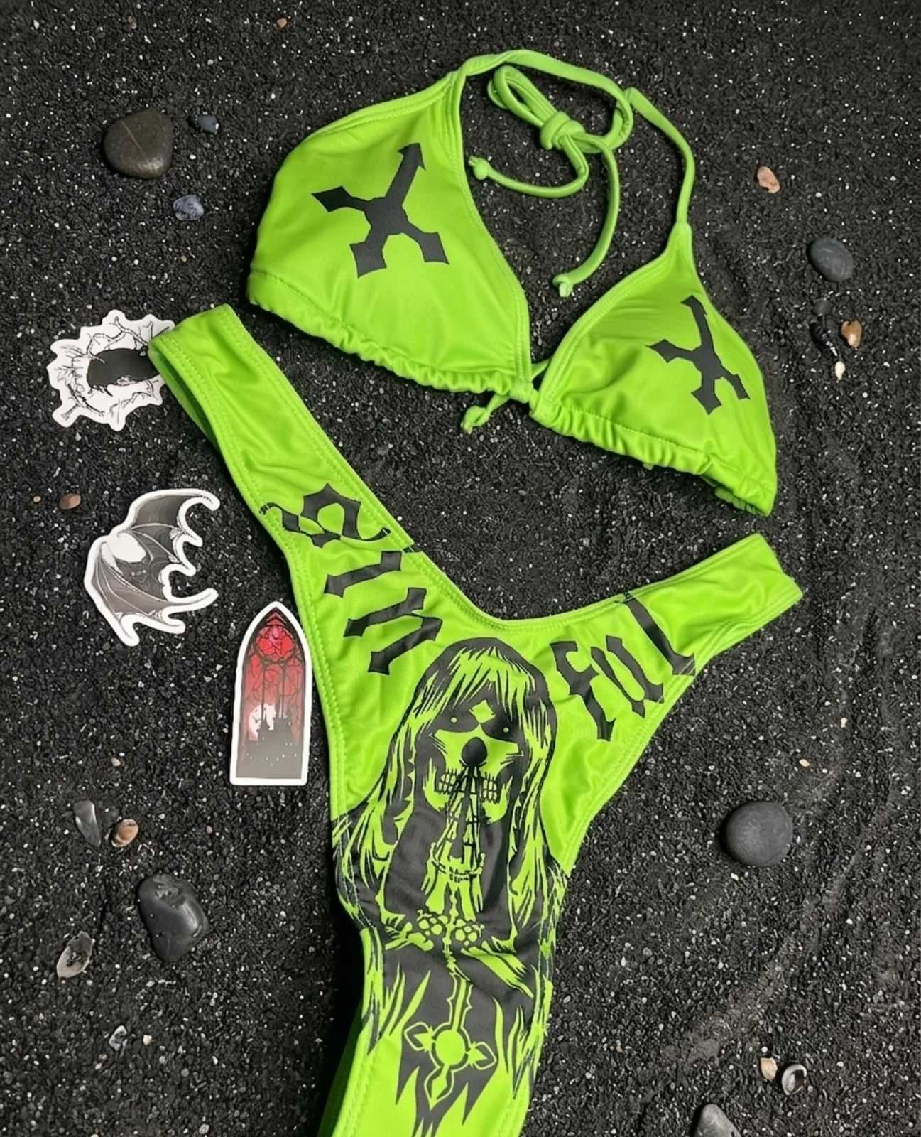 Bikini verde neón con cruces negras en el top y la palabra «sinful» y una parca estampadas en el bottom, sobre arena volcánica negra con stickers góticos