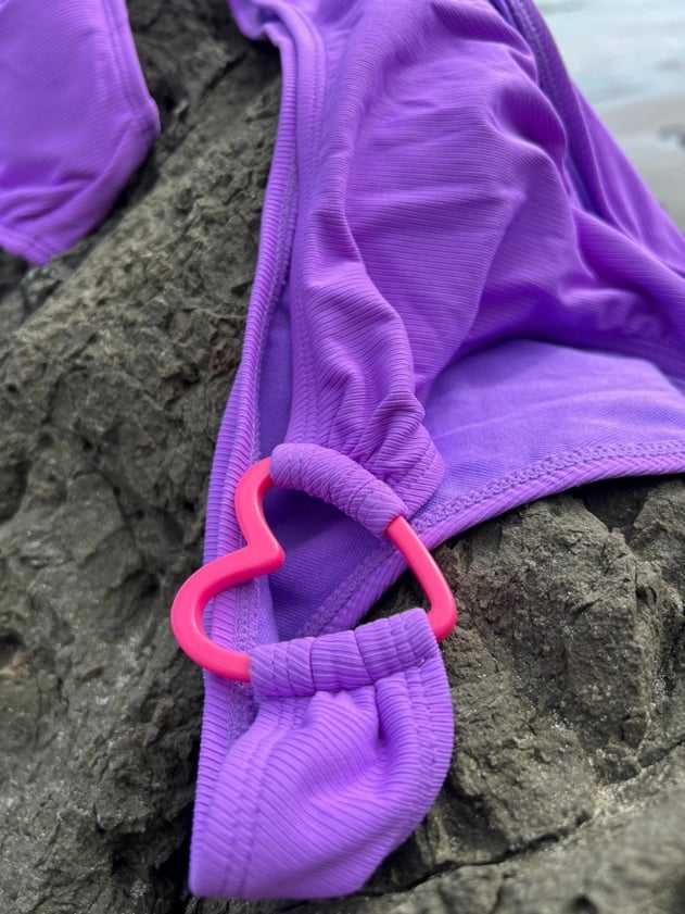 Detalle de un bikini morado con un aro rosado en forma de corazón, sobre una roca