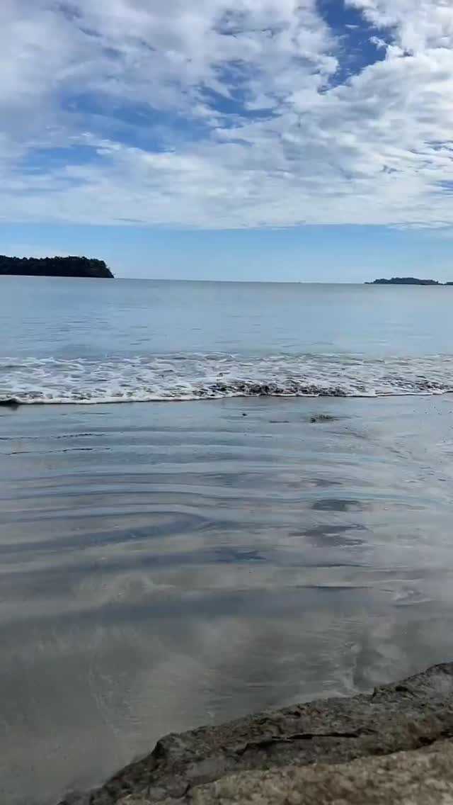 Orilla de una playa de arena oscura con olas suaves, cielo con nubes e islas en el horizonte