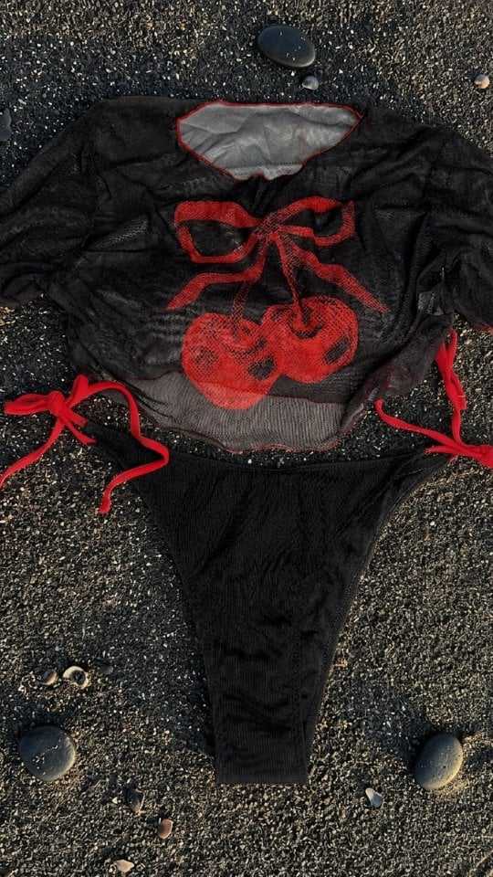 Top de malla negra con cerezas y lazo rojos y bottom negro con tiras rojas, sobre arena de playa